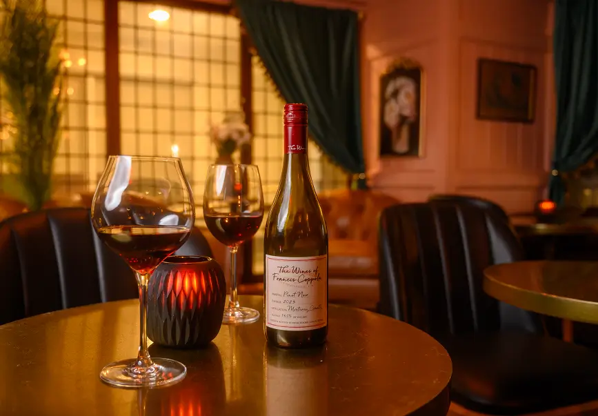 Francis Coppola Pinot Noir 2023 with two glasses on a candlelit table inside Rockwell & Rose, Philadelphia – plush seating and soft pink walls in the background.