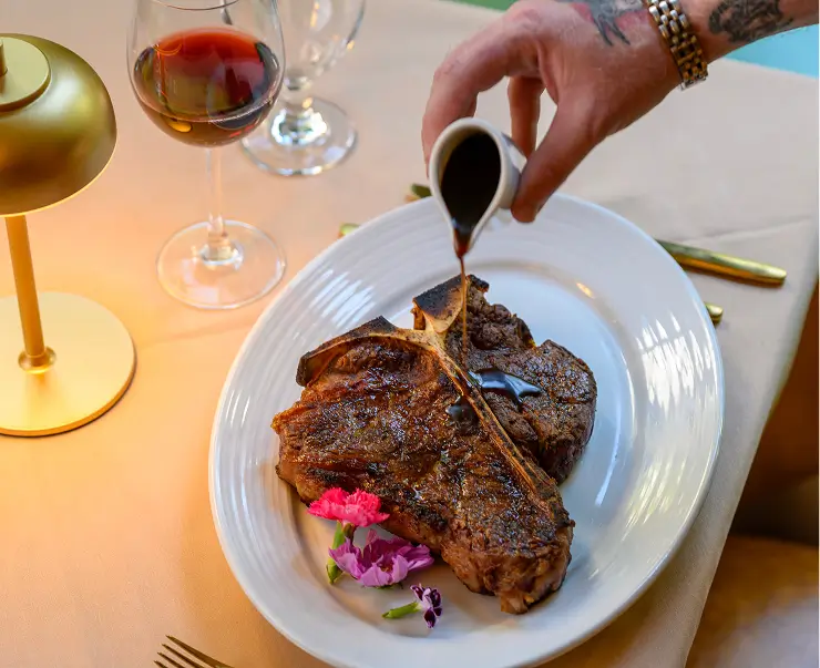 Tableside pour over a dry-aged bone-in ribeye at Rockwell & Rose – served with red wine and floral garnishes in the Rockwell Room.